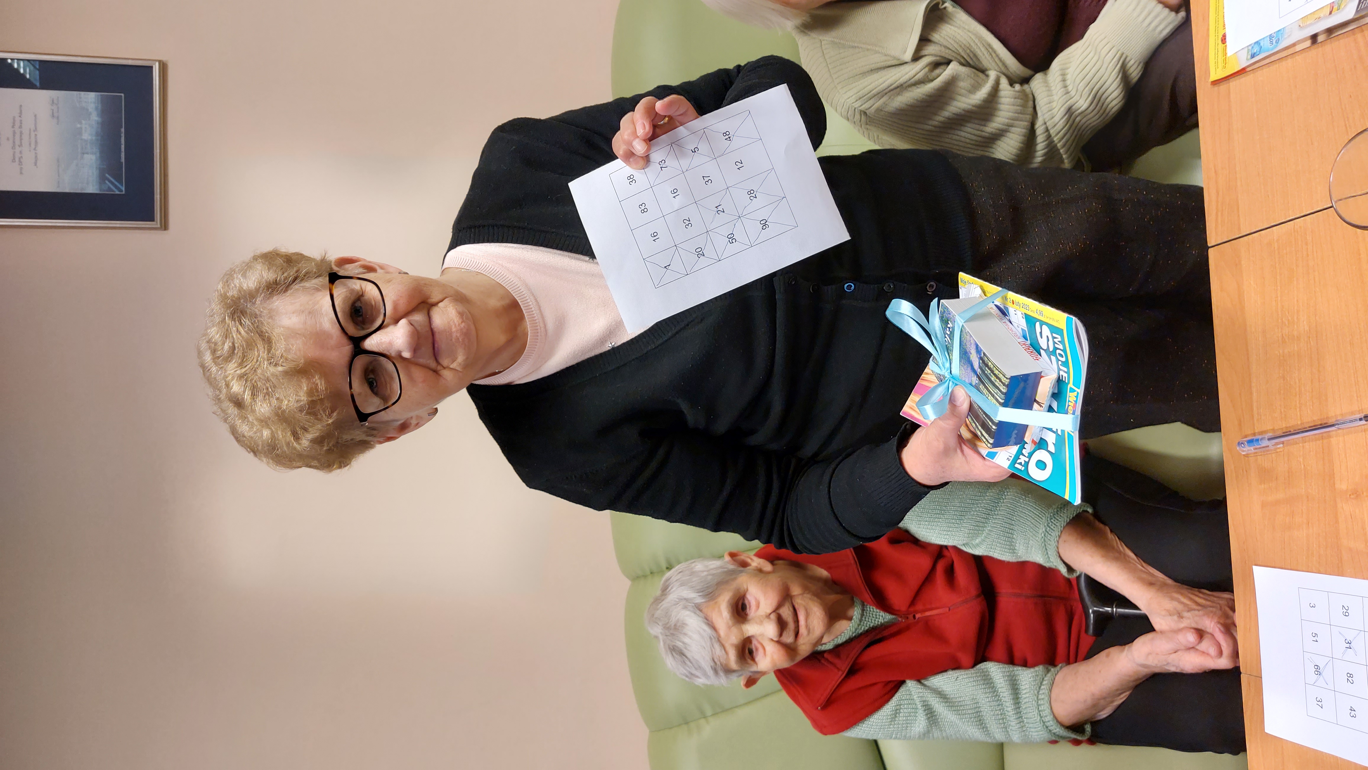 Seniorka trzyma w rękach kartkę do gry w Bingo oraz prezencik