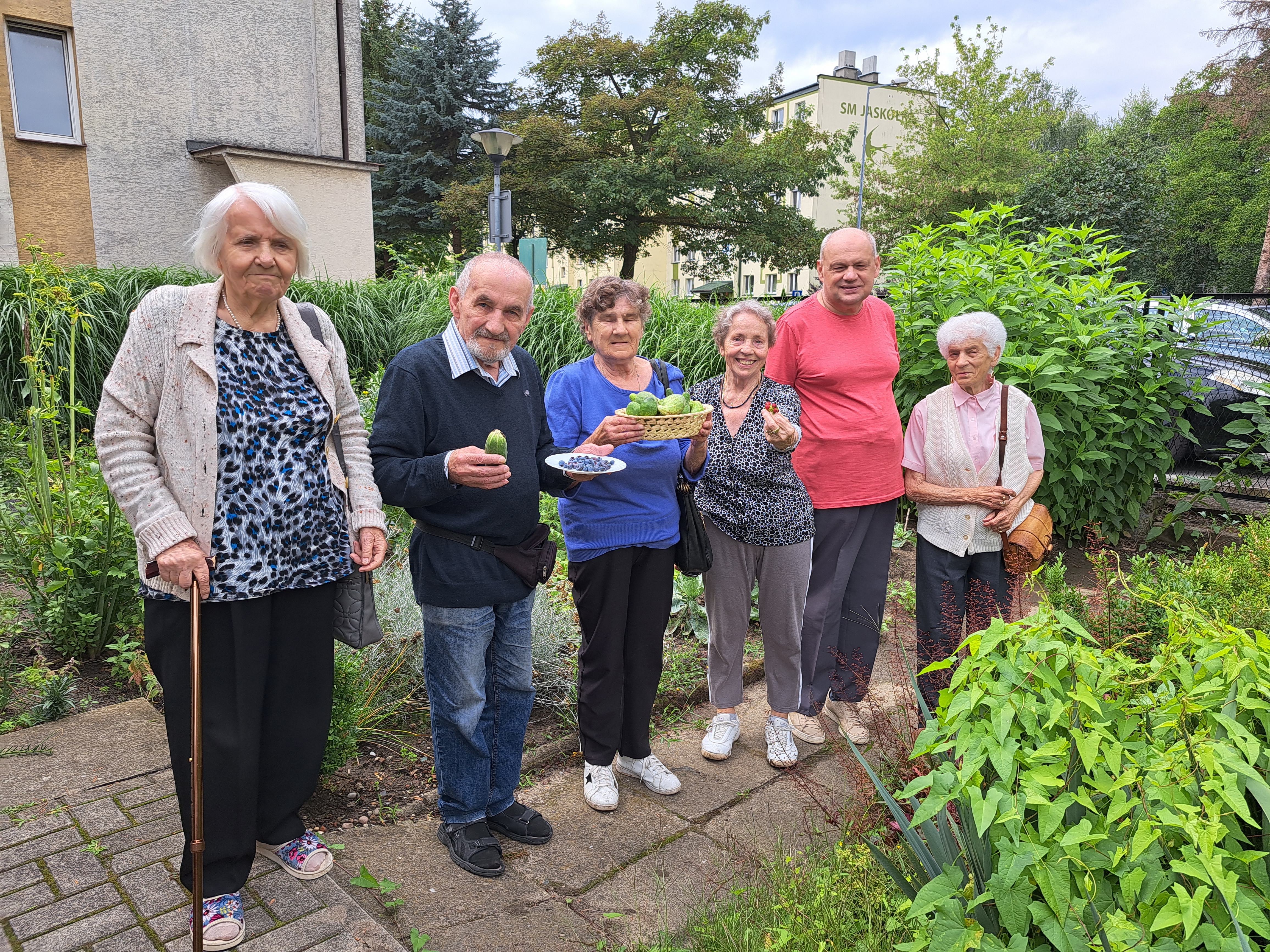 Seniorzy pozują na tle Dziennego Domu z zebranymi owocami i warzywami