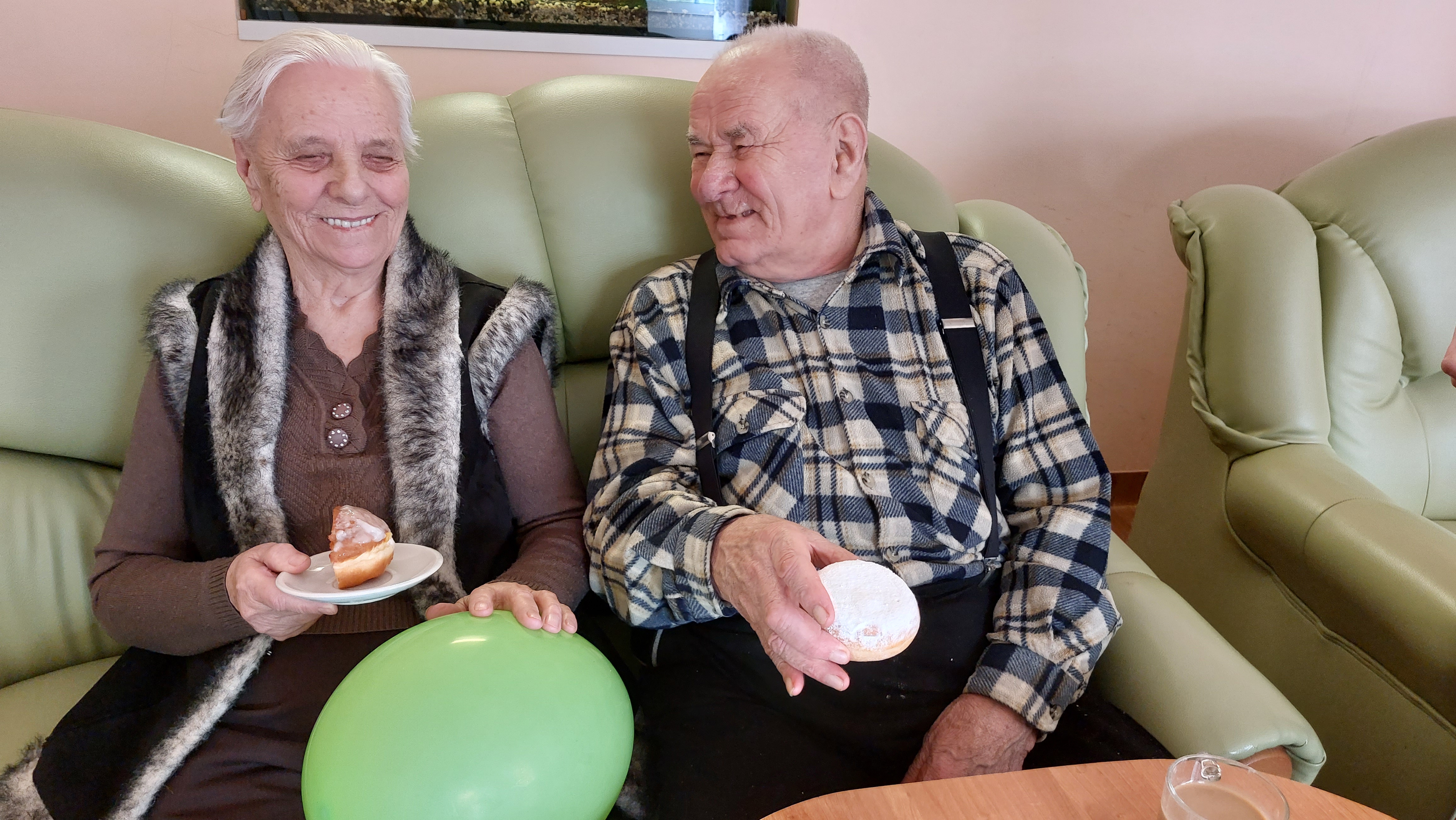 Seniorka oraz Senior siedzą na kanapie i jedzą pączki