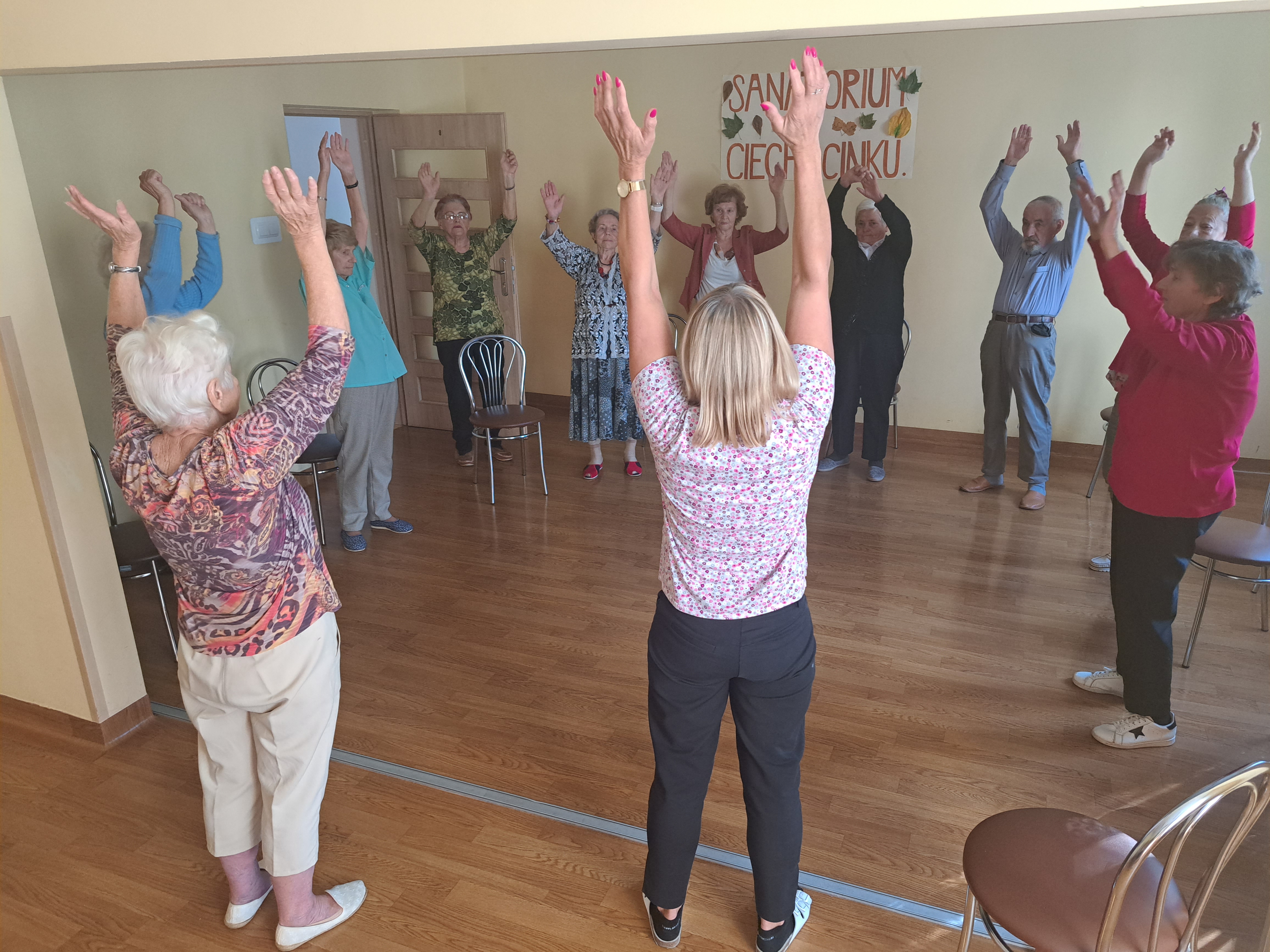 Seniorzy stoją w kółku z wyciągniętymi rękoma w górze, rozciągają się.