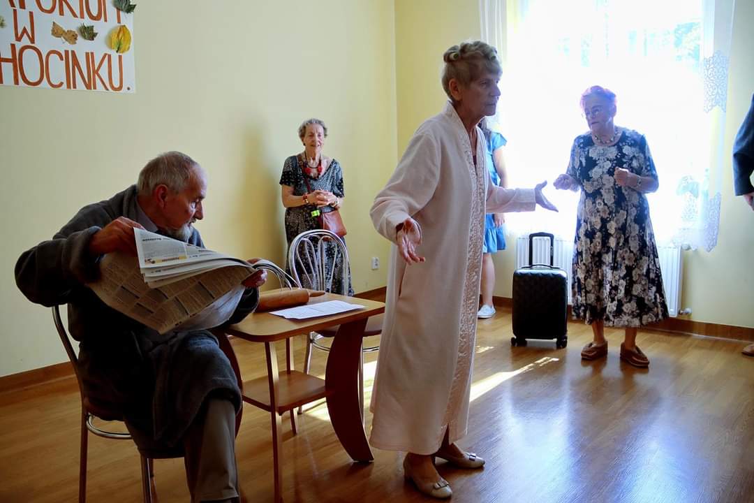 Seniorzy wykonują skecz Sanatorium w Ciechocinku, w tle siedzi Senior z gazetą, na pierwszym planie Seniorka w szlafroku deklamuje swoją rolę