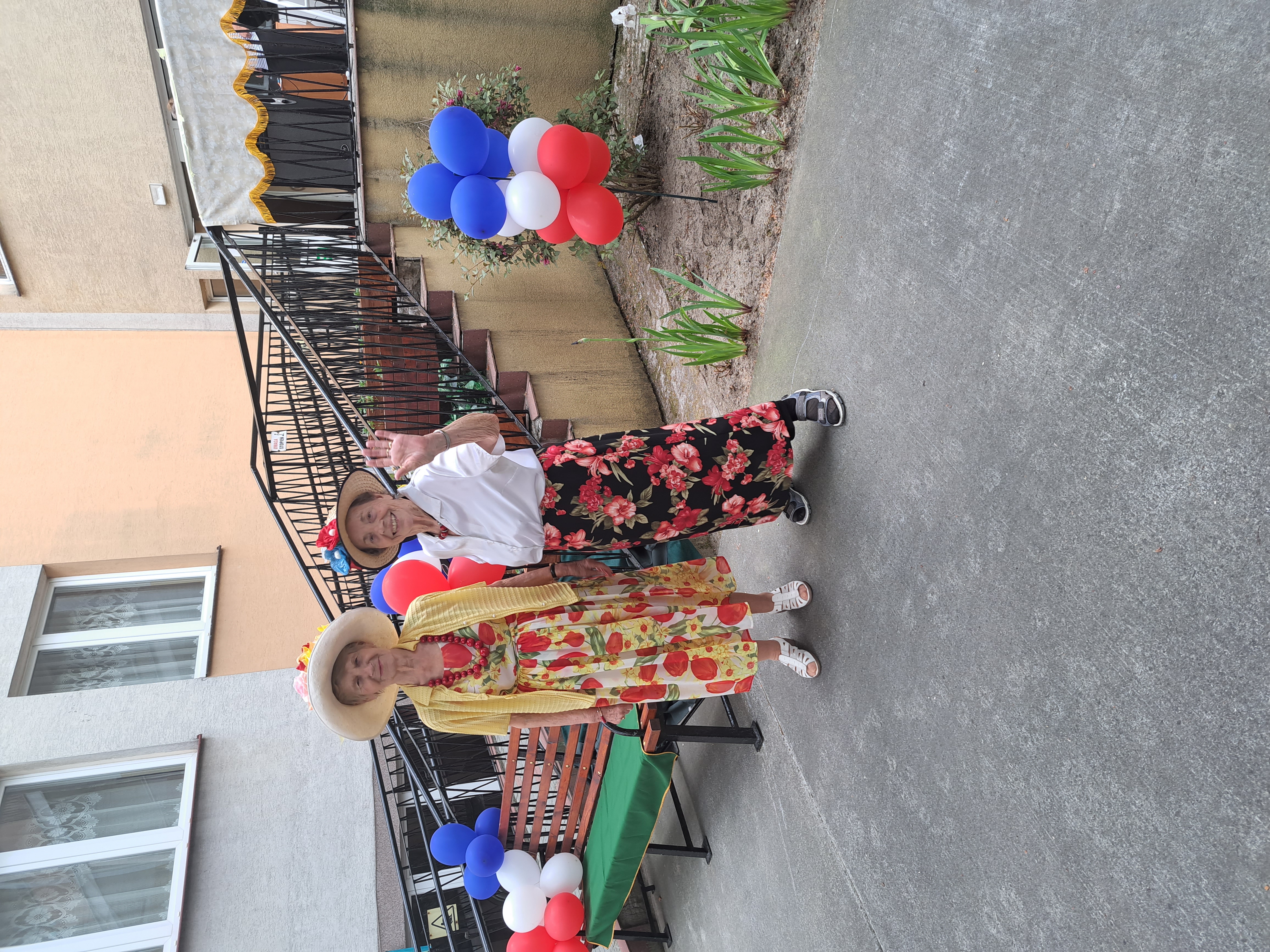 Dwie Seniorki w kolorowych spódnicach oraz kapeluszach pozują do zdjęcia w ogrodzie Domu