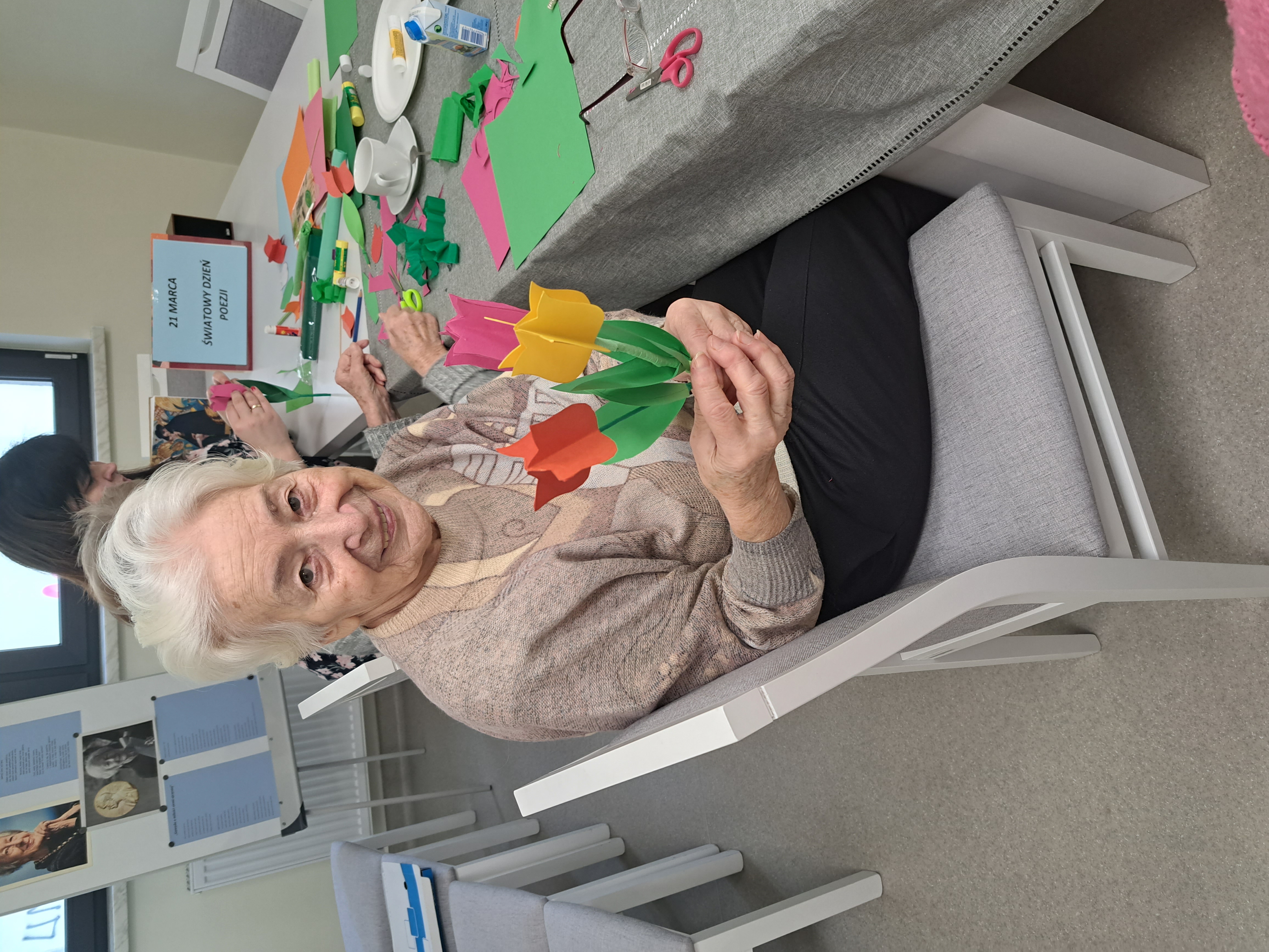 Seniorka siedzi przy stole, pozuje do zdjęcia z bukiecikiem własnoręcznie zrobionych tulipanów z papieru