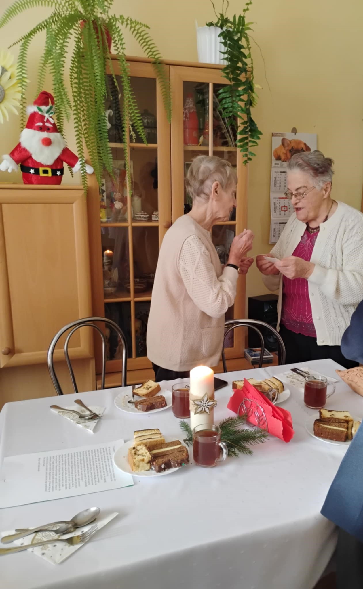 Dwie Seniorki dzielą się opłatkiem, na pierwszym planie znajduje się zastawiony stół z jedzeniem