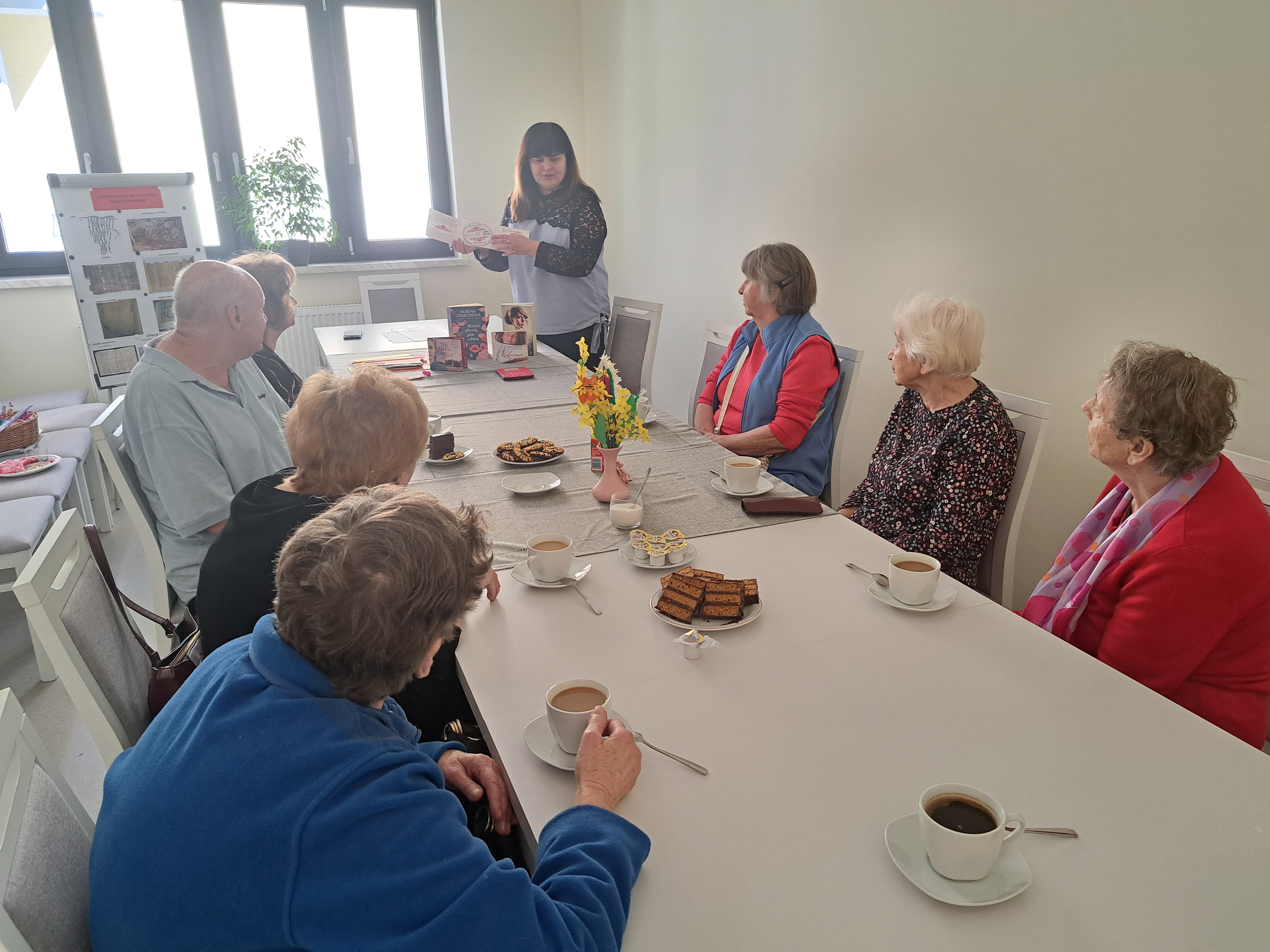 Seniorzy siedzą przy stołe, przy kawie i ciasteczkach, słuchają opowiadania pracowniczki biblioteki