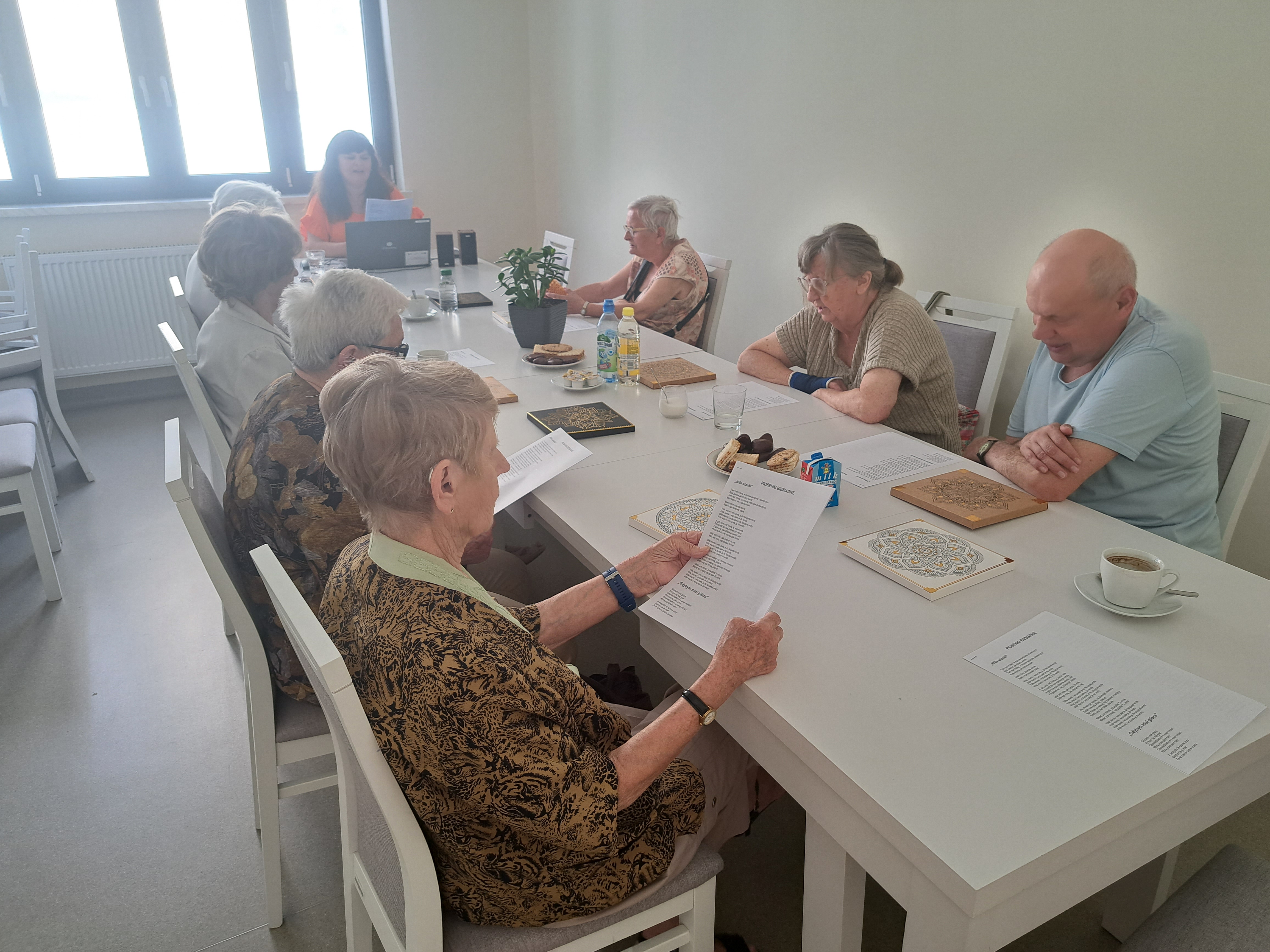 Seniorzy siedzą przy długim stole, na jego końcu siedzi Pani opowiadająca o życiu Jana Szczepanika