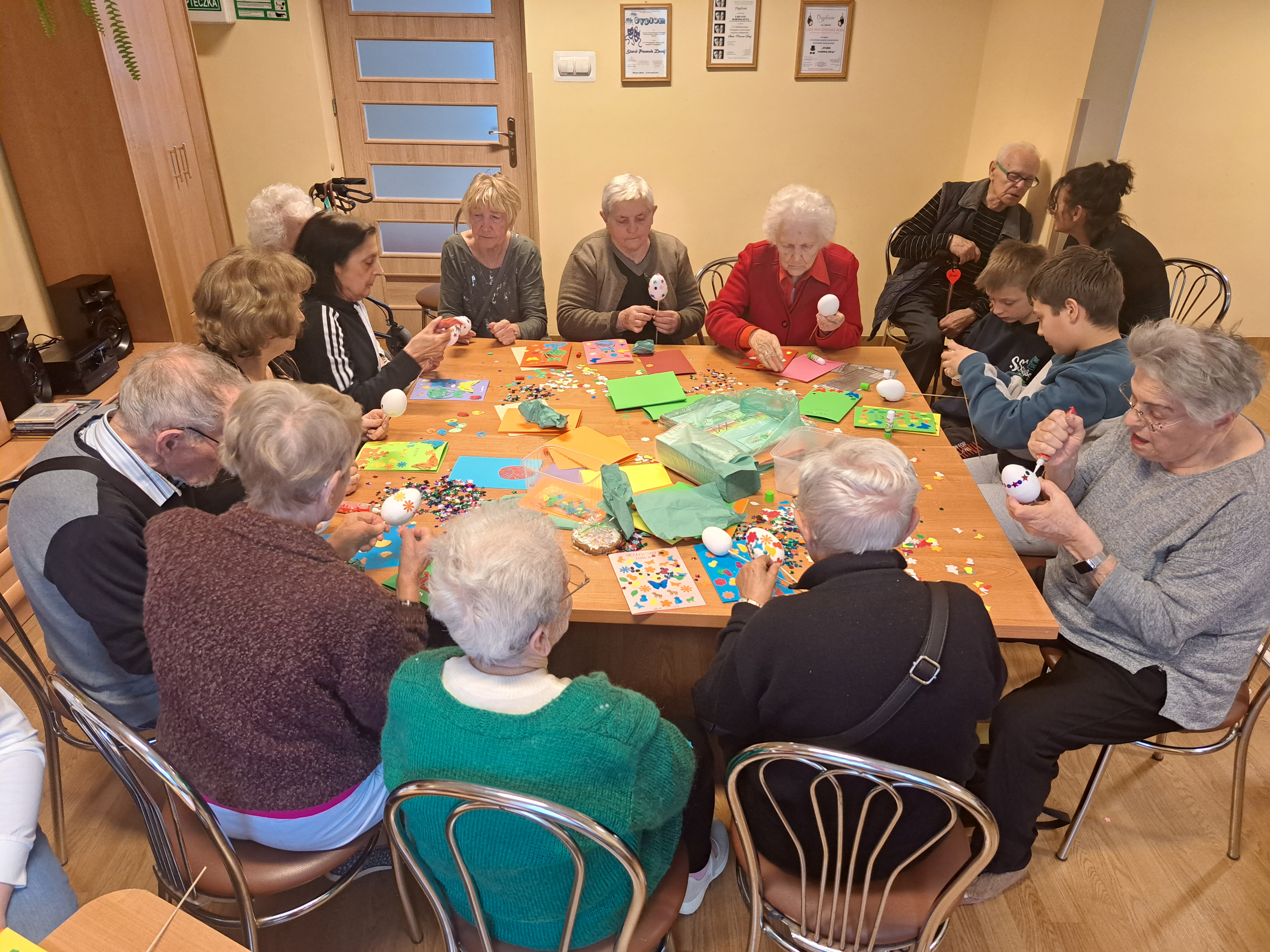 Seniorzy siedzą przy stole z dziećmi i wykonują razem robótki z kolorowego papieru