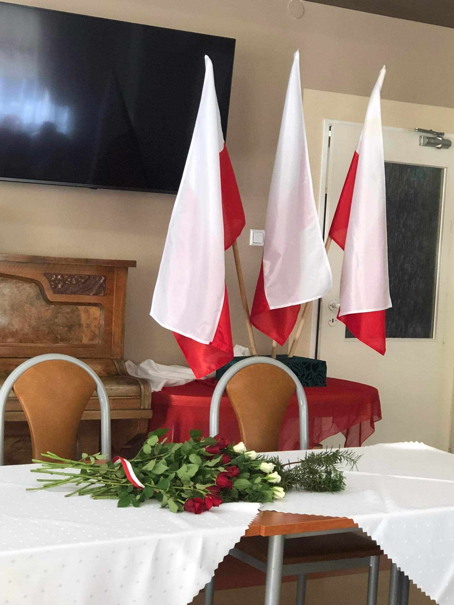 W tle trzy flagi polski, z przodu stół z białym obrusem, na którym leżą róże