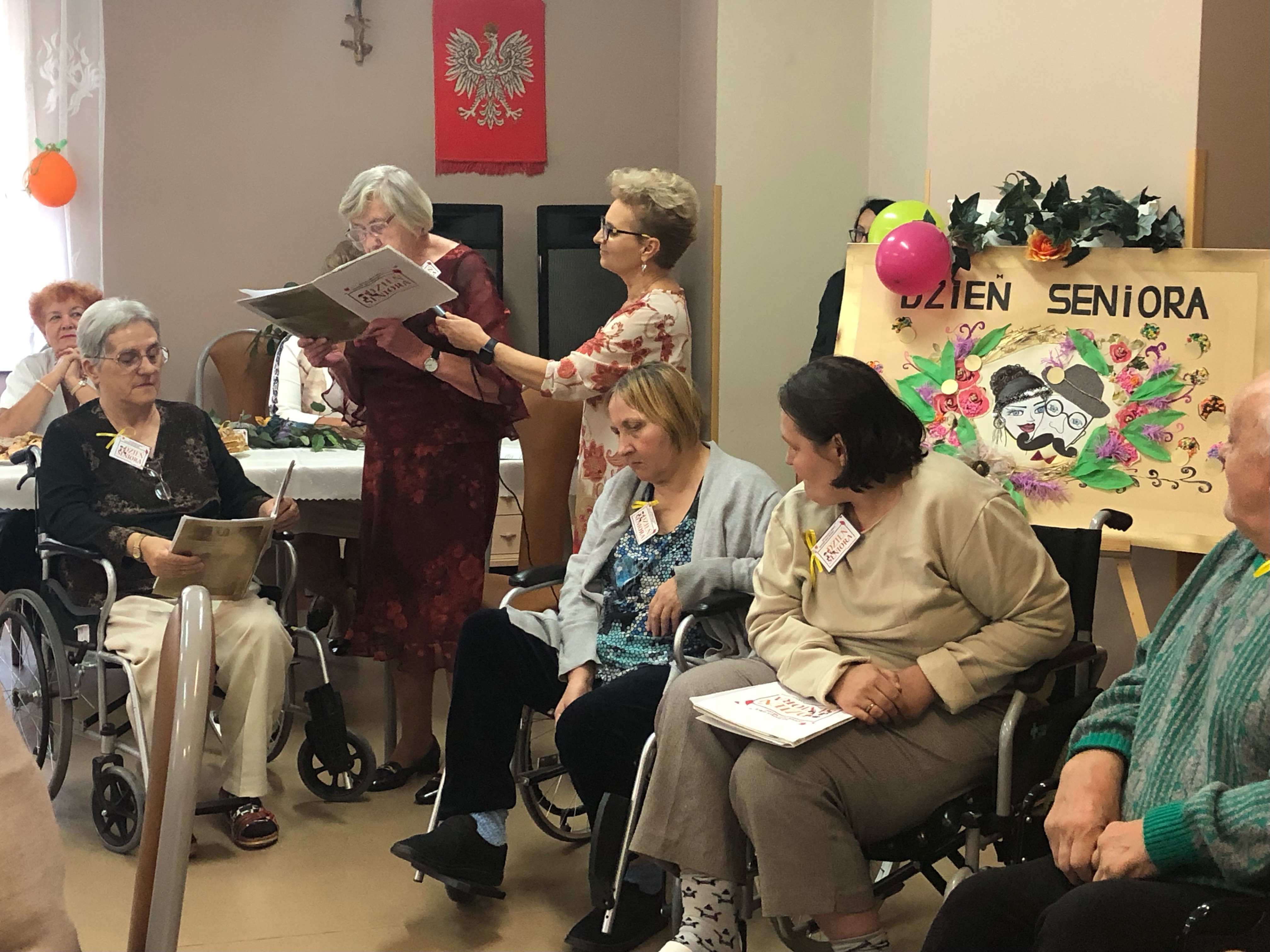 Czworo seniorów siedzi na wózkach inwalidzkich,jedna Seniorka deklamuje wiersz 