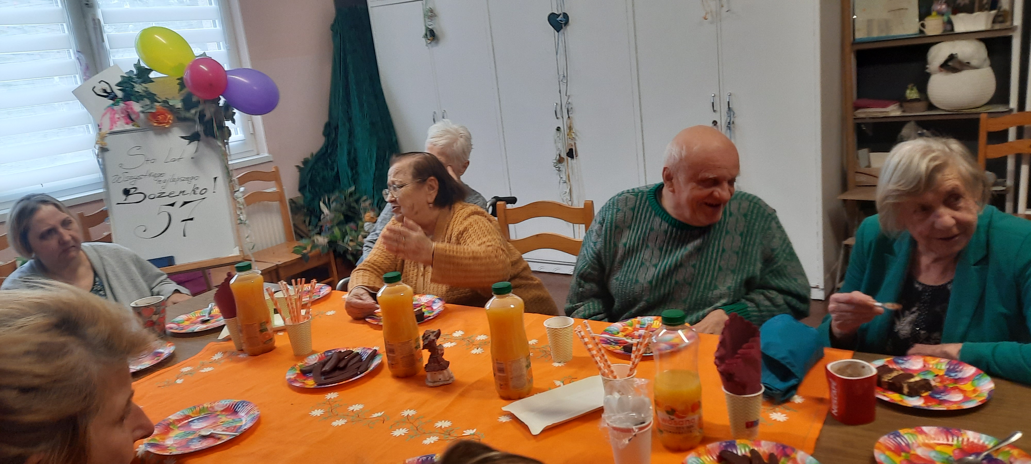 Grupa seniorów siedzi przy stole, rozmawia oraz je tort