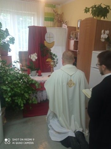 Dwaj kapłani klęczą przed ołtarzem przyozdobionym czerwono białą wstęgą z namalowanym kielichem