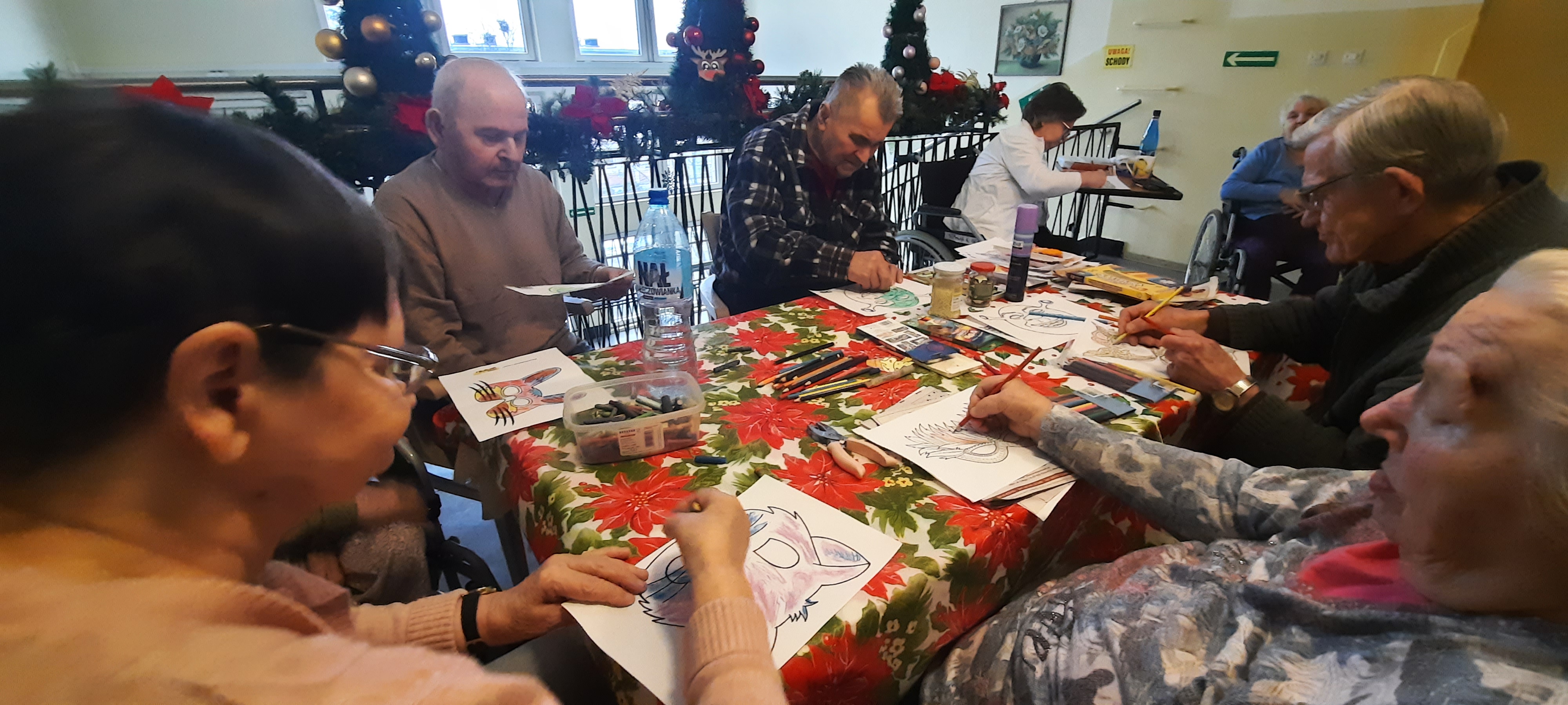 Grupa Seniorów siedzi przy stole, kolorują maski karnawałowe