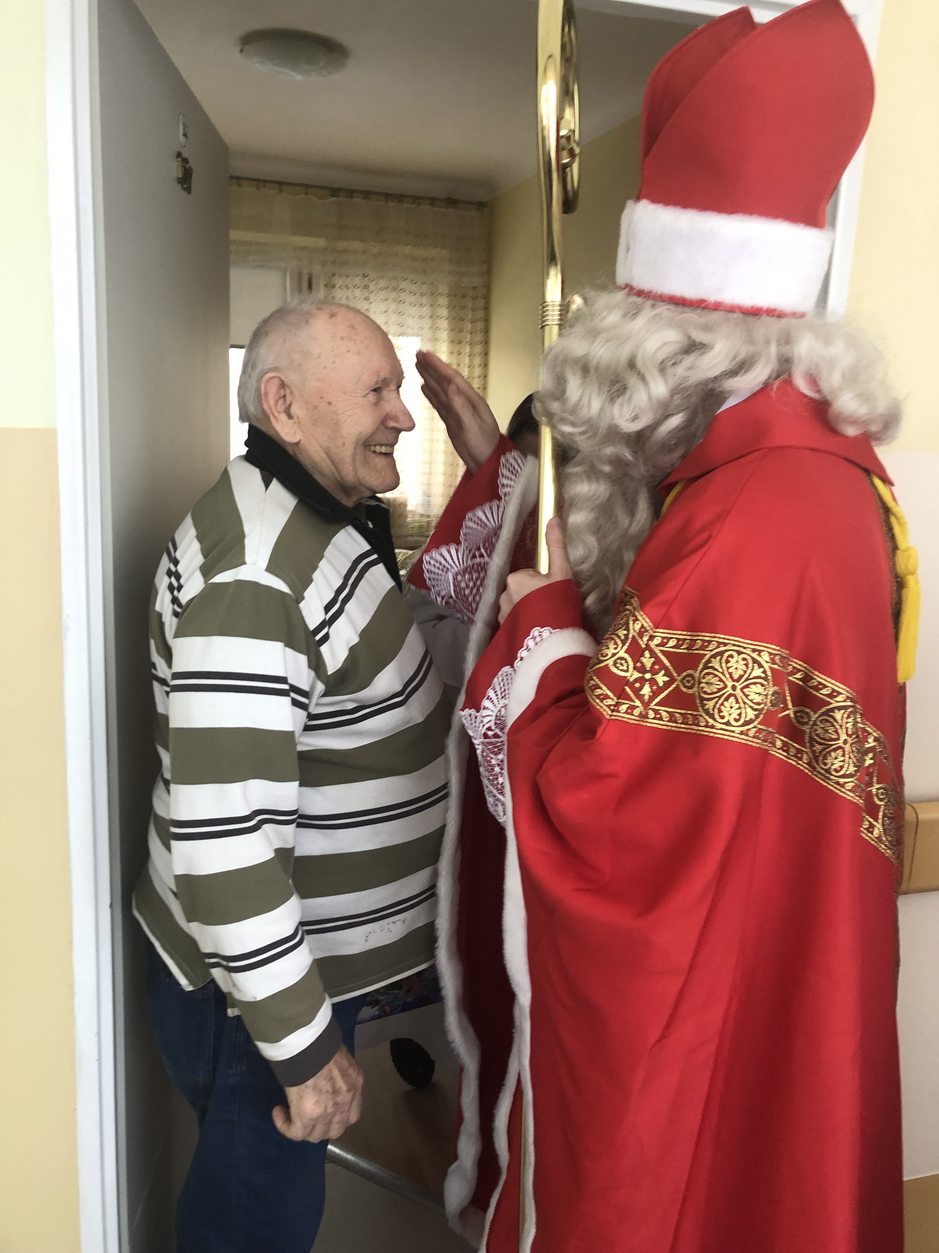 Mikołaj oraz Senior stoją w progu pokoju, Mikołaj wykonuje znak krzyża na głowie Seniora