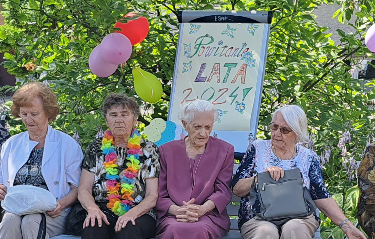 Seniorzy siedzą w ogrodzie, w tle stoi tablica z napisem Powitanie lata