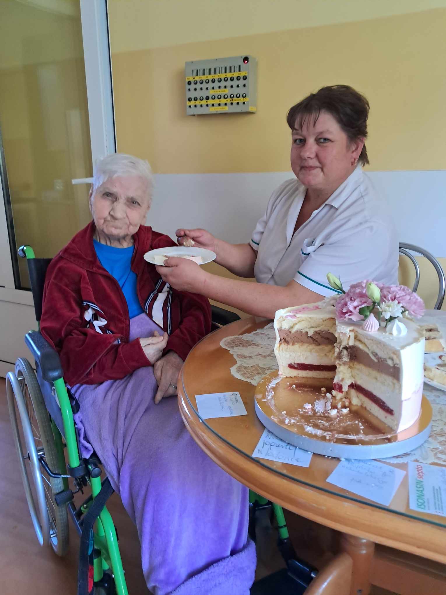 Pracownica DPS siedzi przy stoliku z Panią Weroniką, pomaga jej w jedzeniu torta