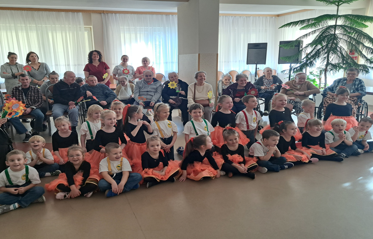 Zdjęcie zrobione na świetlicy Domu Seniorzy siedzą na krzesłach z tyłu,a z przodu na podłodze po turecku siedzą dzieci