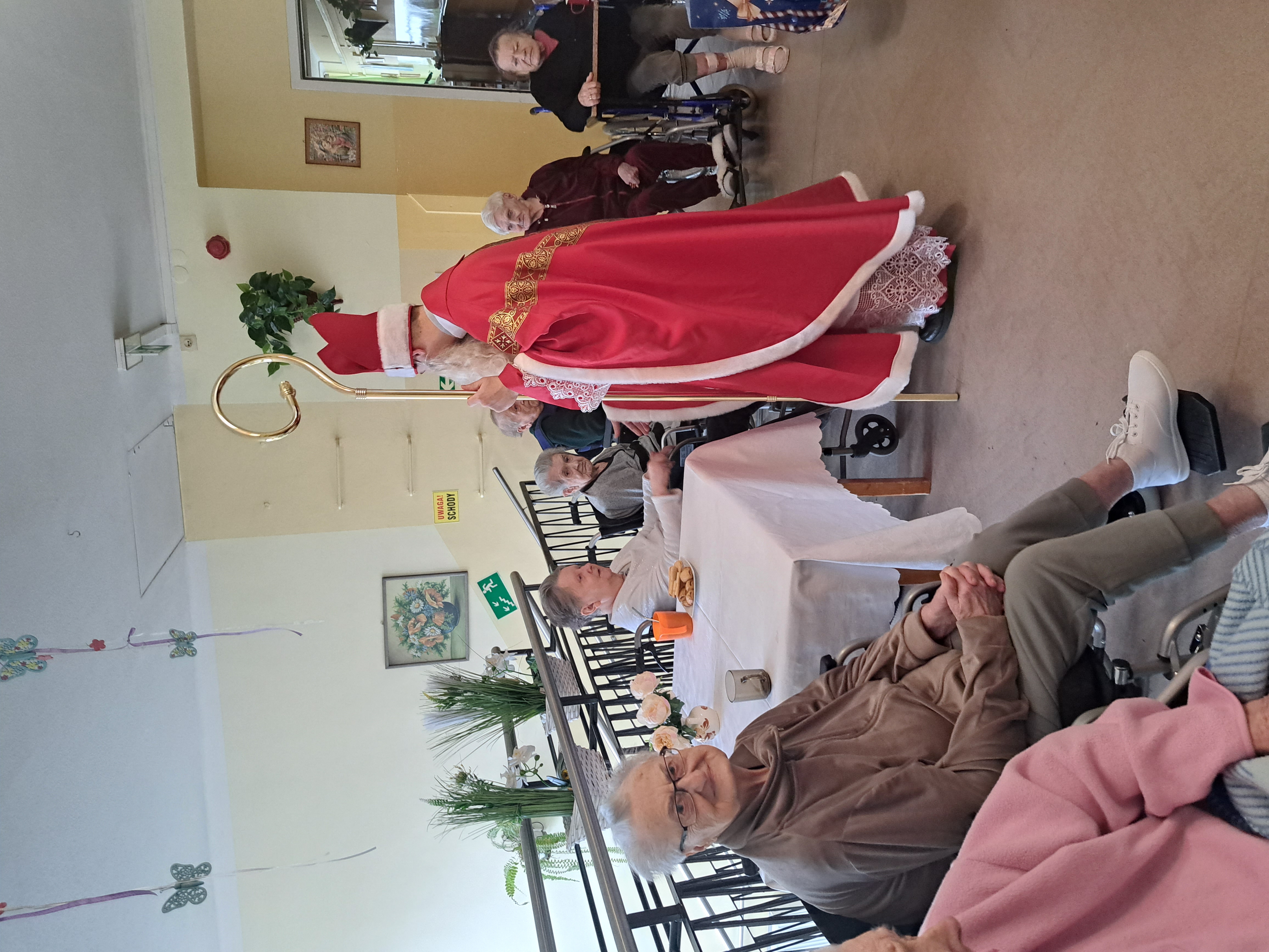 Mikołaj wita się z Seniorami siedzącymi przy stoliku z kawą i ciasteczkami