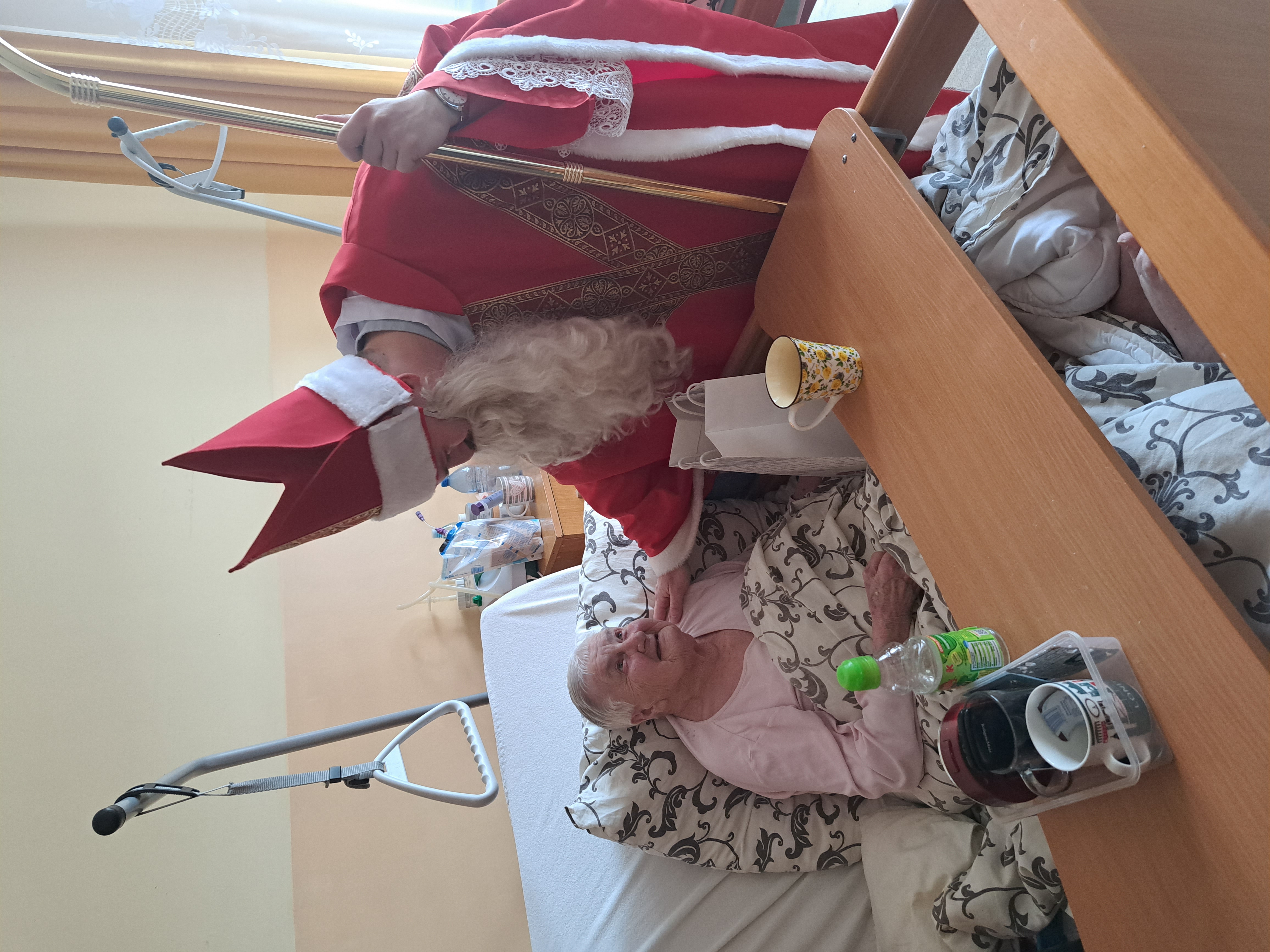 Seniorka leży na łóżku, Mikołaj pochyla się nad nią i składa życzenia