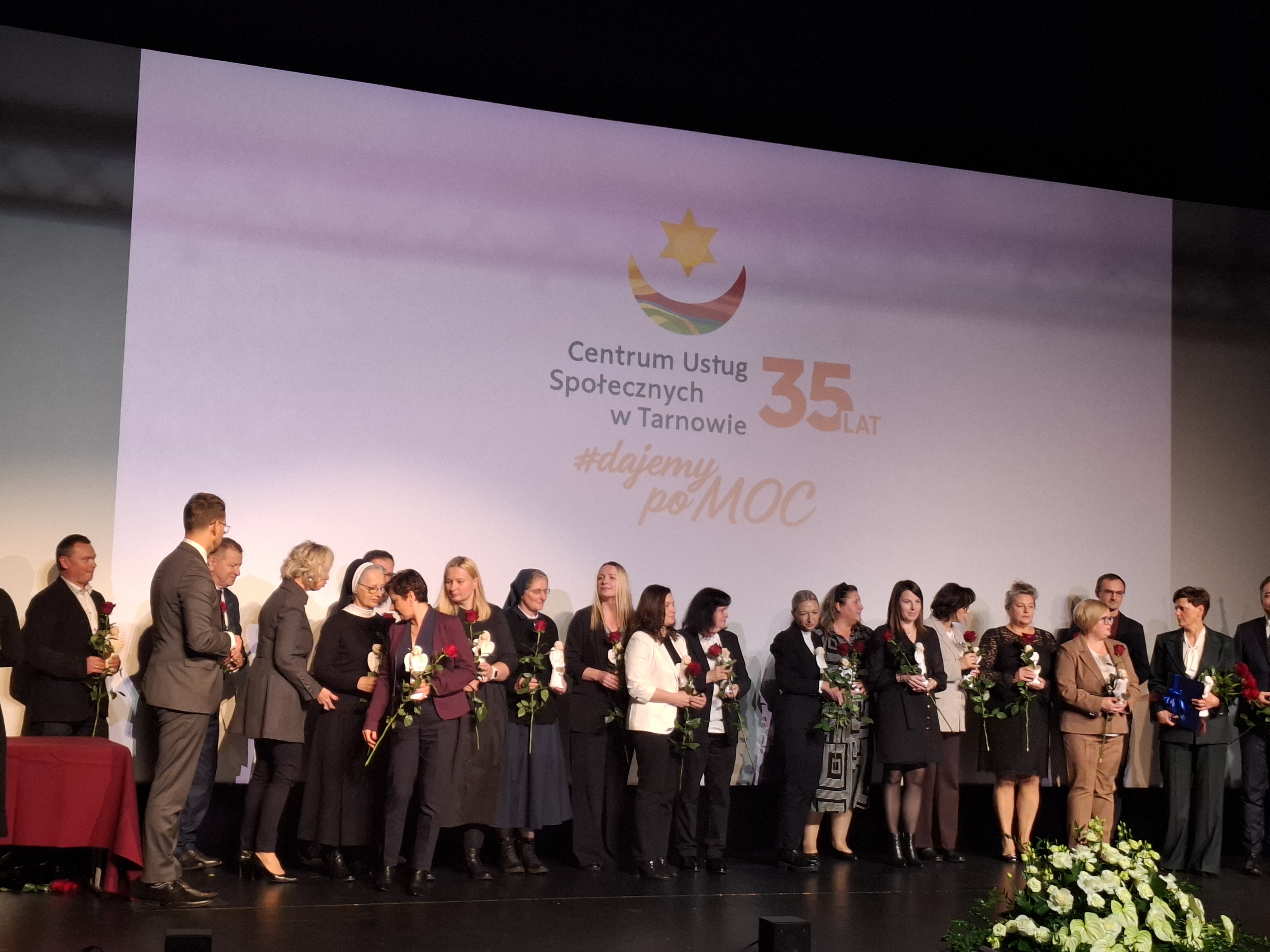 Szeroki kadr na grupę elegancko ubranych ludzi stoi na scenie, trzymajac róże w ręku, za nimi znajduje wyświetlony napis Centrum Usług Wspólnych w Tarnowie 35 lat dajemy PoMOC