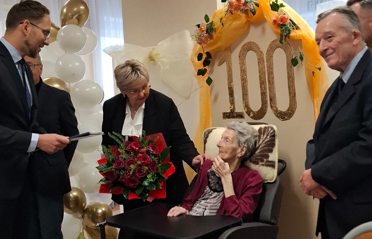 Pani Zosia siedzi na tle standu z liczbą 100, dokoła niej dwoje mężczyzn i kobieta wręczająca Seniorce kwiaty