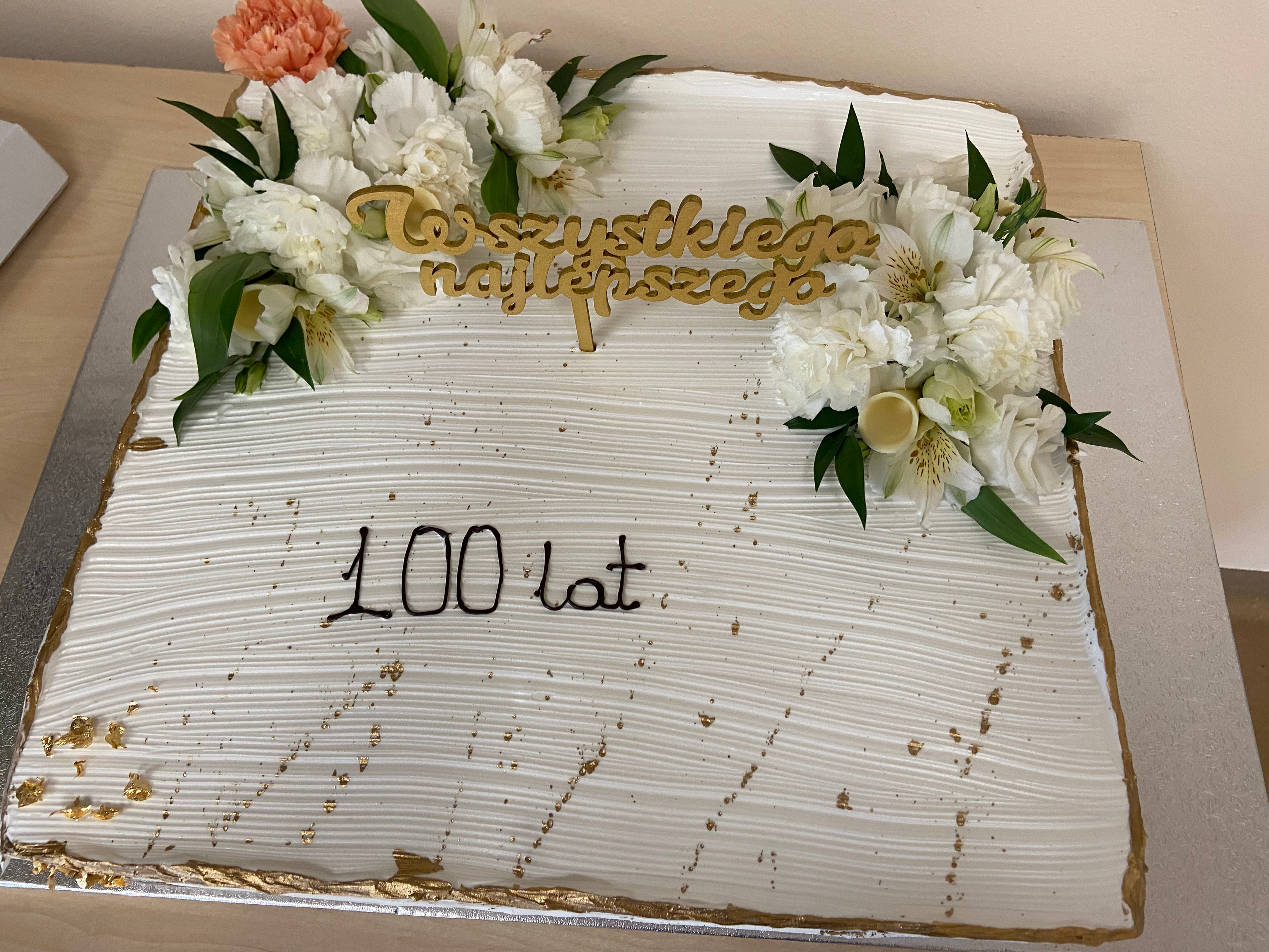 Biały tort ozdobiony kwiatami, z napisem 100 lat