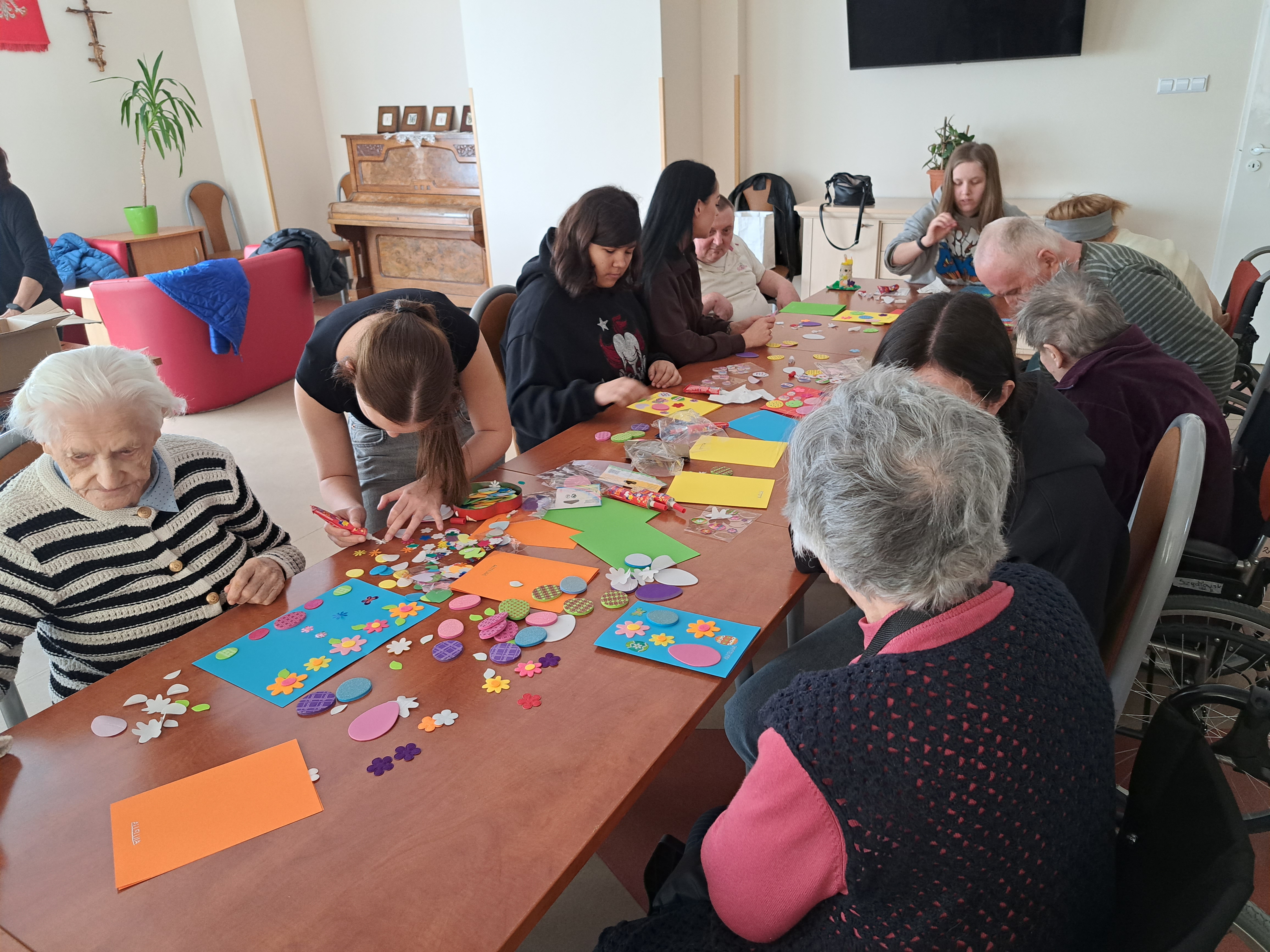 Seniorzy siedzą przy stole, na którym leży papier kolorowy, z którego wykonują kartki wielkanocne
