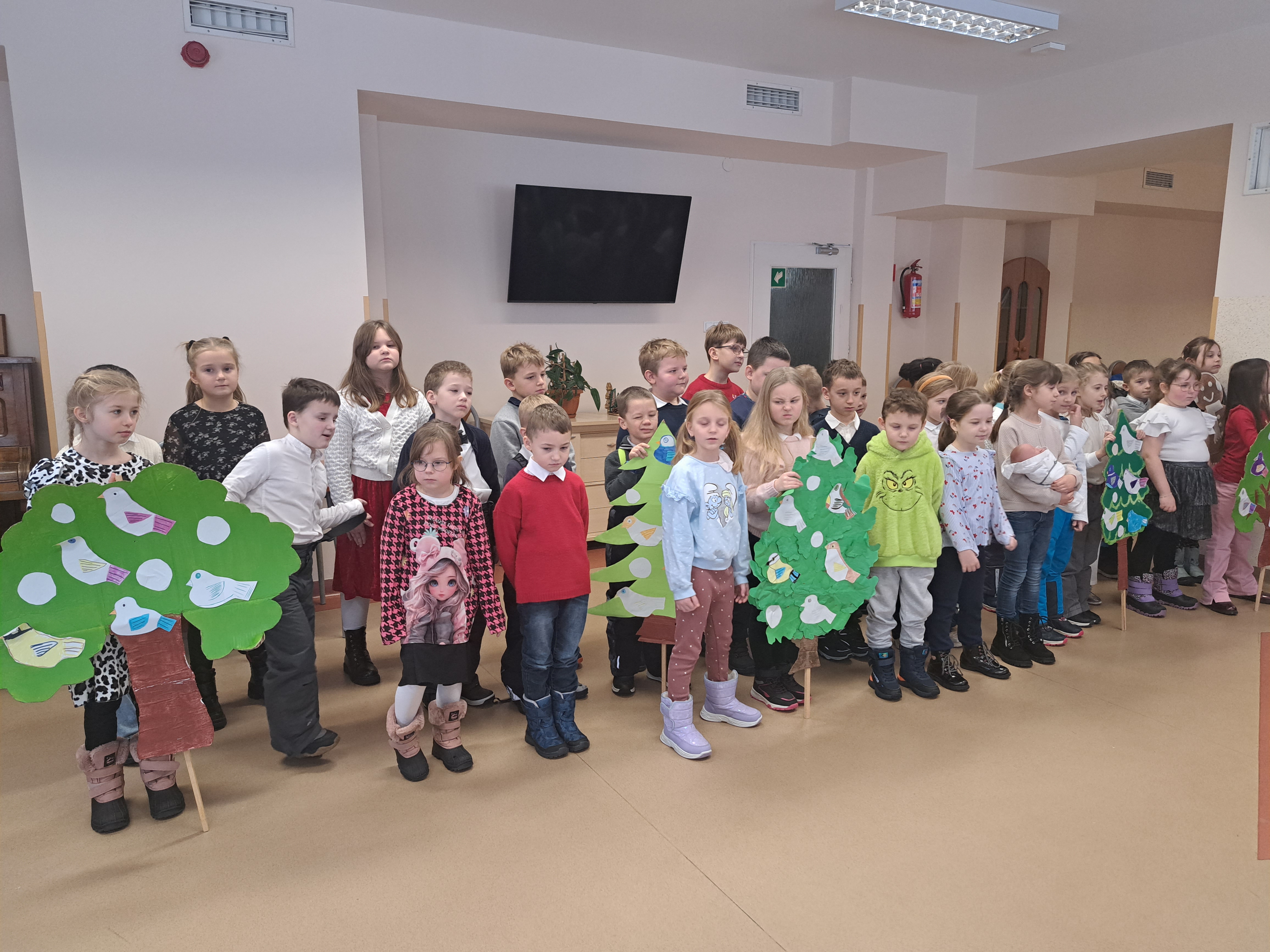 Grupa dzieci występuje przed Seniorami, niektóre trzymają makiety drzew w rękach