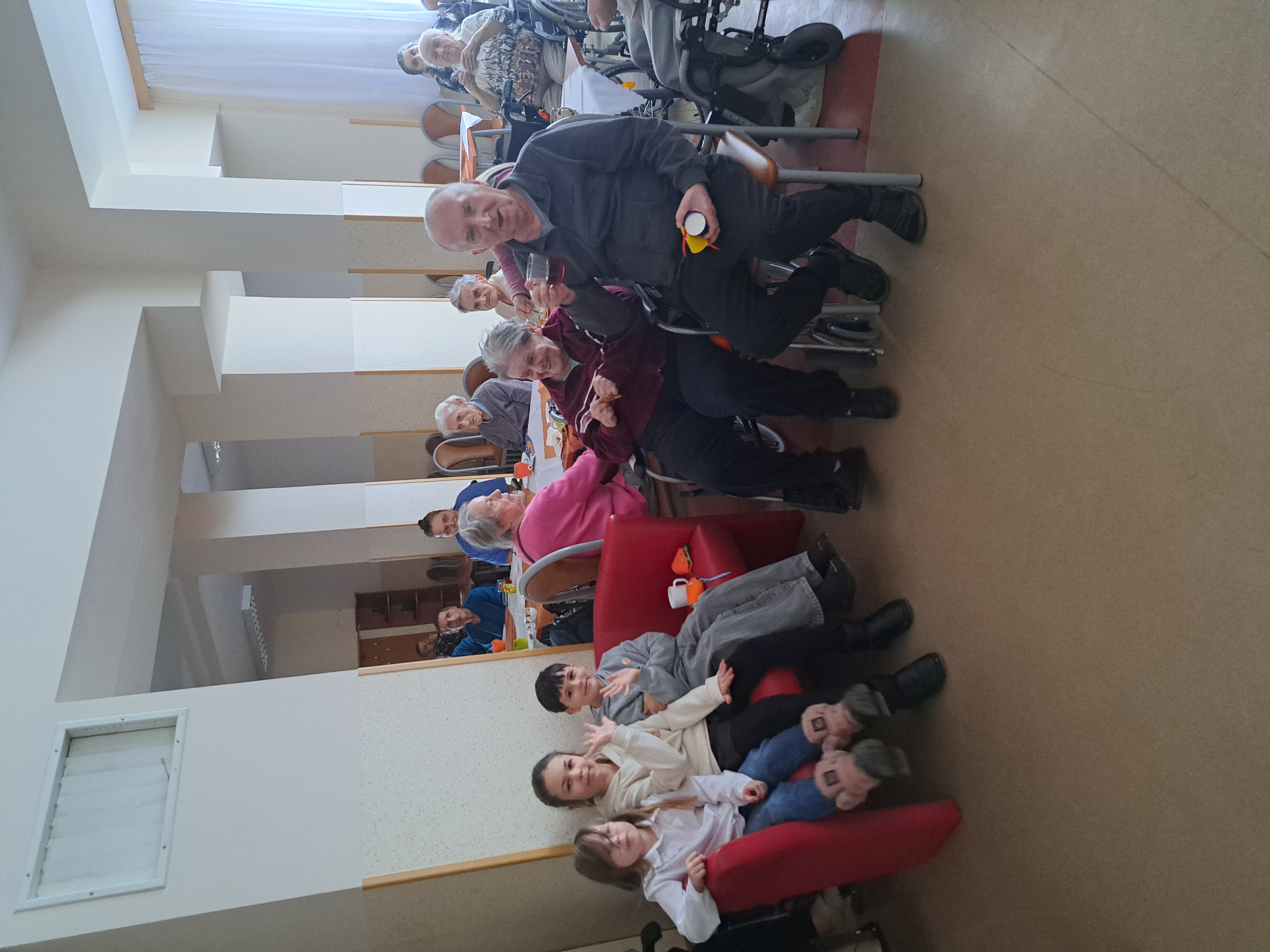 Na pierwszym planie Seniorzy uśmiechają się do zdjęcia, obok siedzą przedszkolaki, w tle Seniorzy kosztują słodki poczęstunek