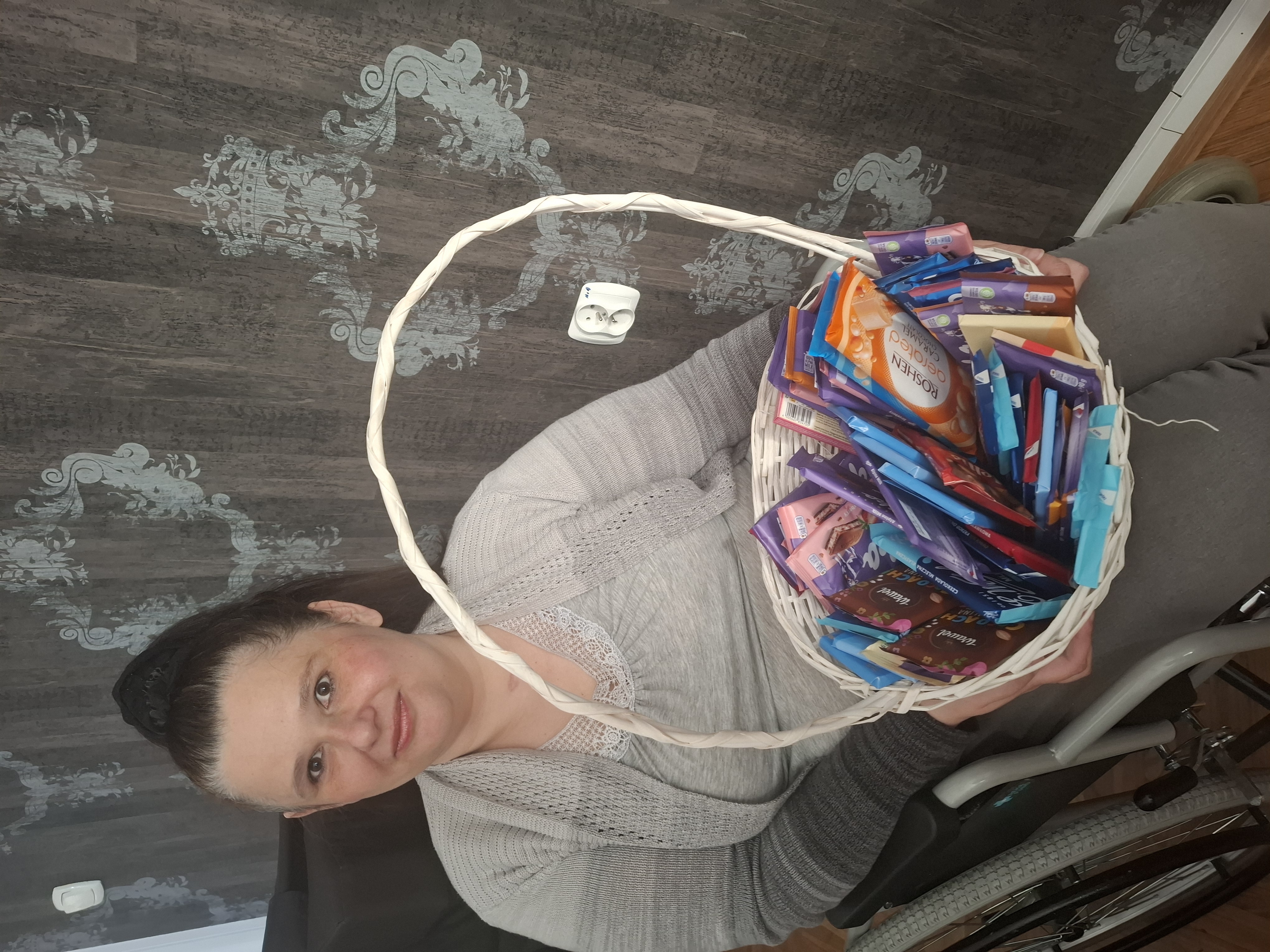 Kobieta siedząca na wózku inwalidzkim trzyma na kolanach duży kosz wypełniony czekoladami