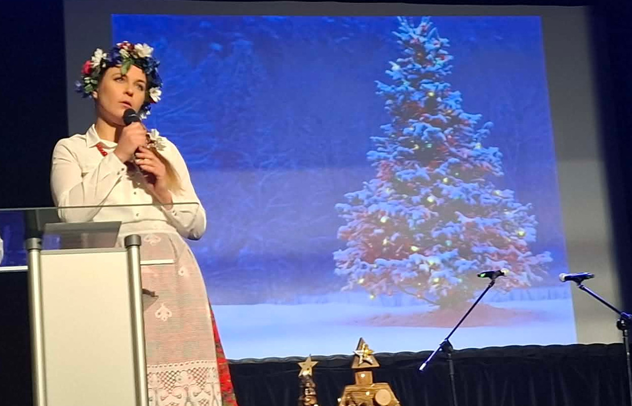 Kobieta w ludowym stroju stoi na scenie, obok niej znajduje się rzutnik z wyświetlonym drzewkiem świątecznym