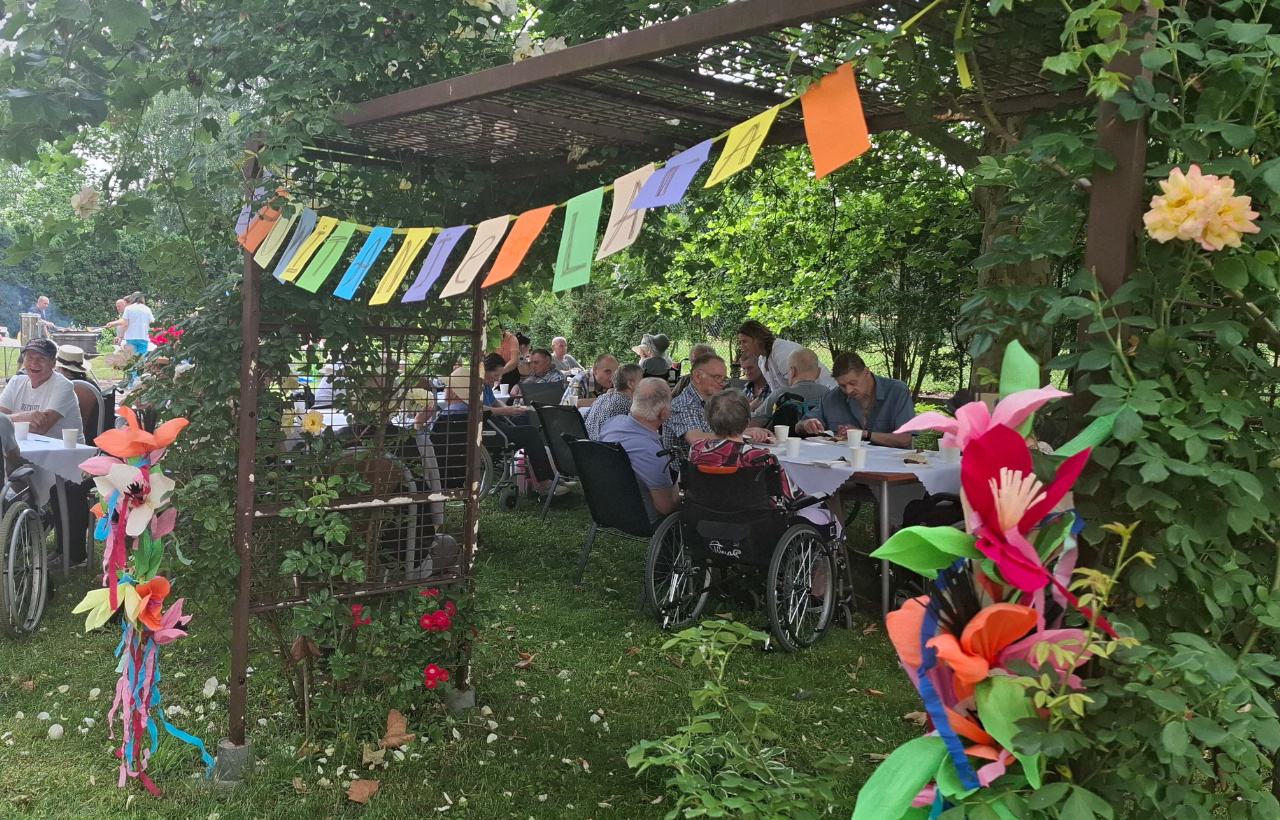 Zdjęcie ogrodu domu, w oddali widać rozłożone stoły, przy których siedzą Seniorzy