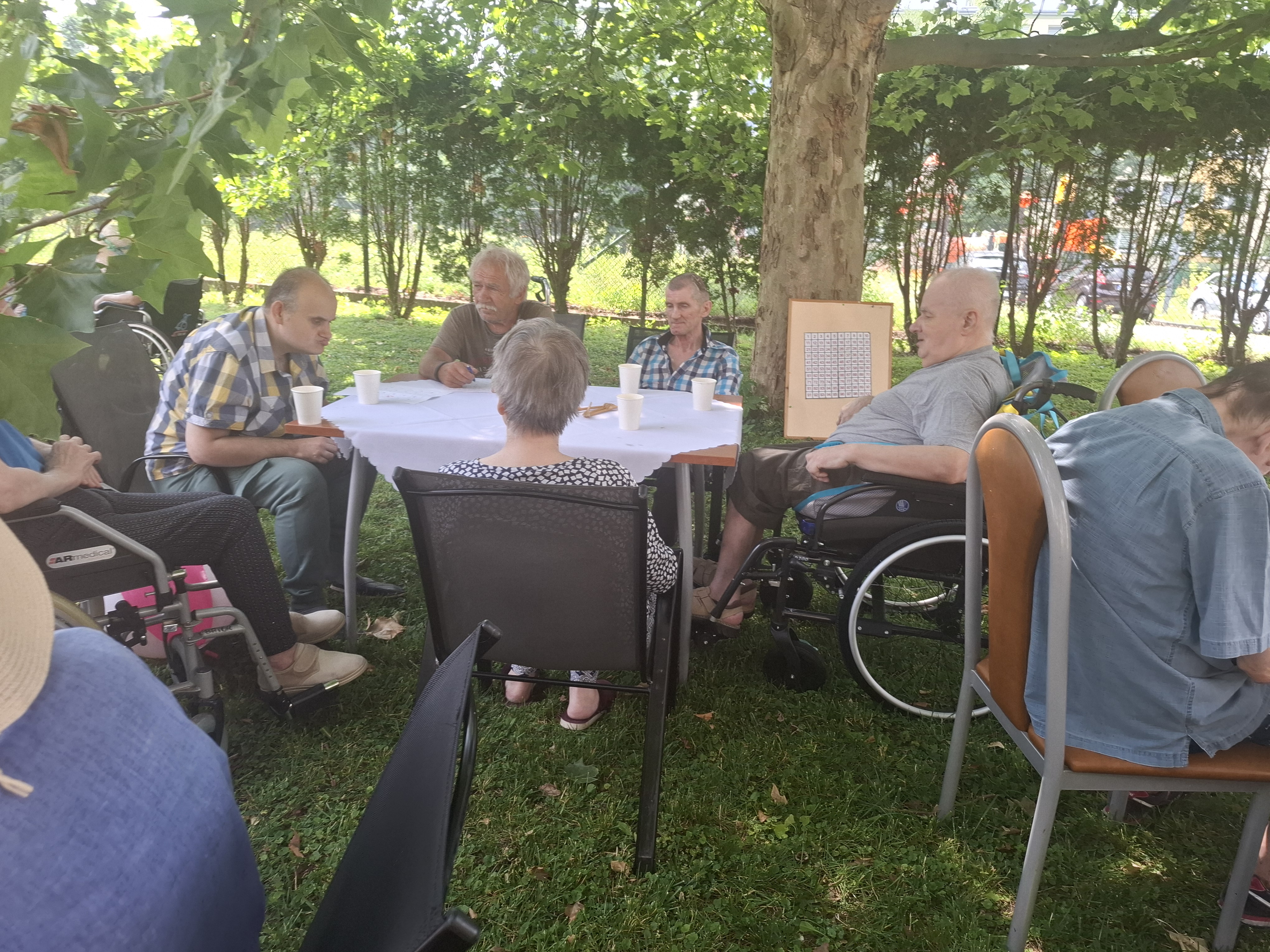 Ujęcie na mały czteroosobowy stolik ustawiony pod drzewem. Przy stoliku siedzą Seniorzy