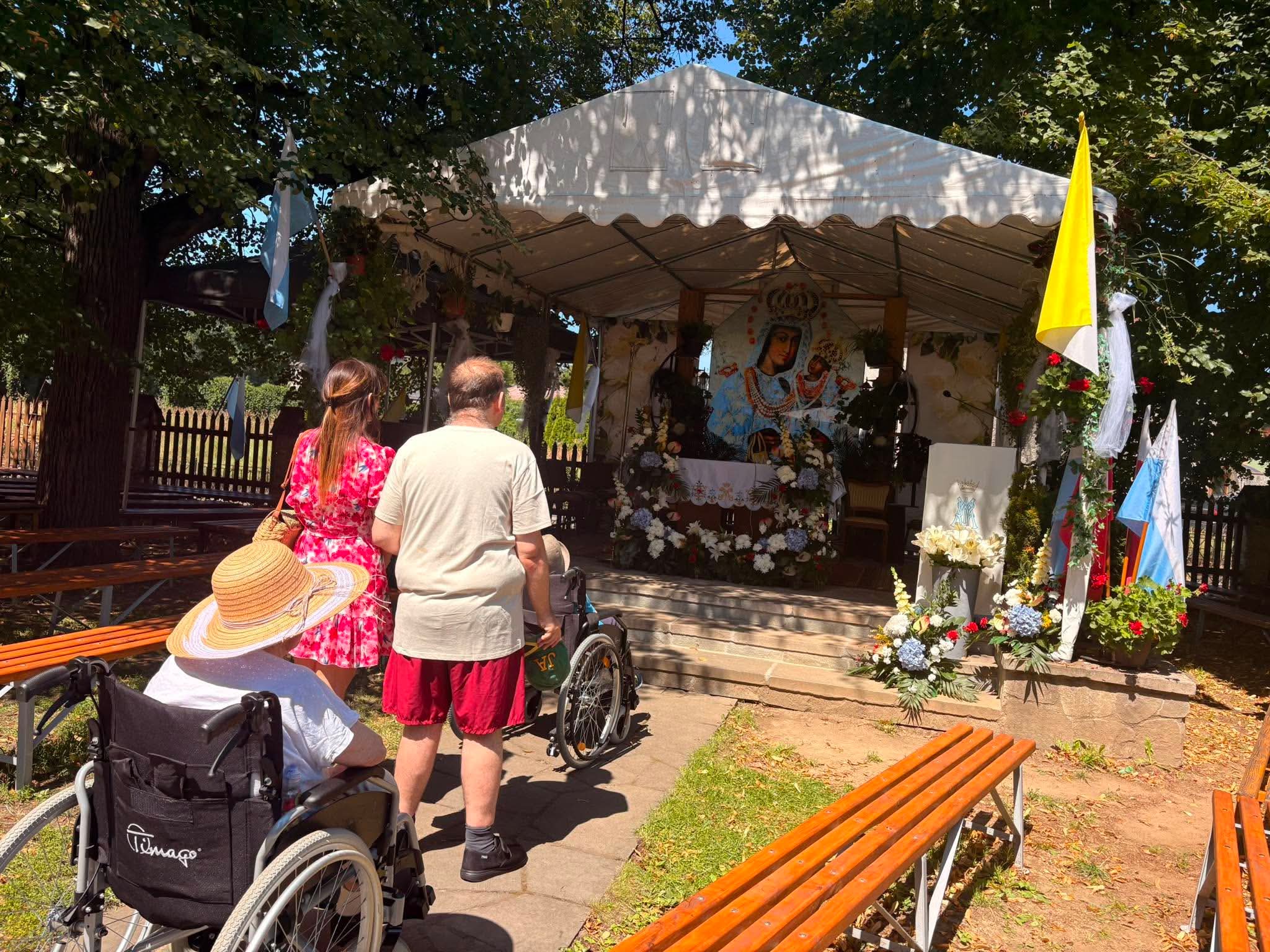 Pracownica Domu oraz Senior i Seniorka na wózku inwalidzkim modlą się przy ołtarzu Matki Boskiej znajdującym się na zewnątrz Kościoła