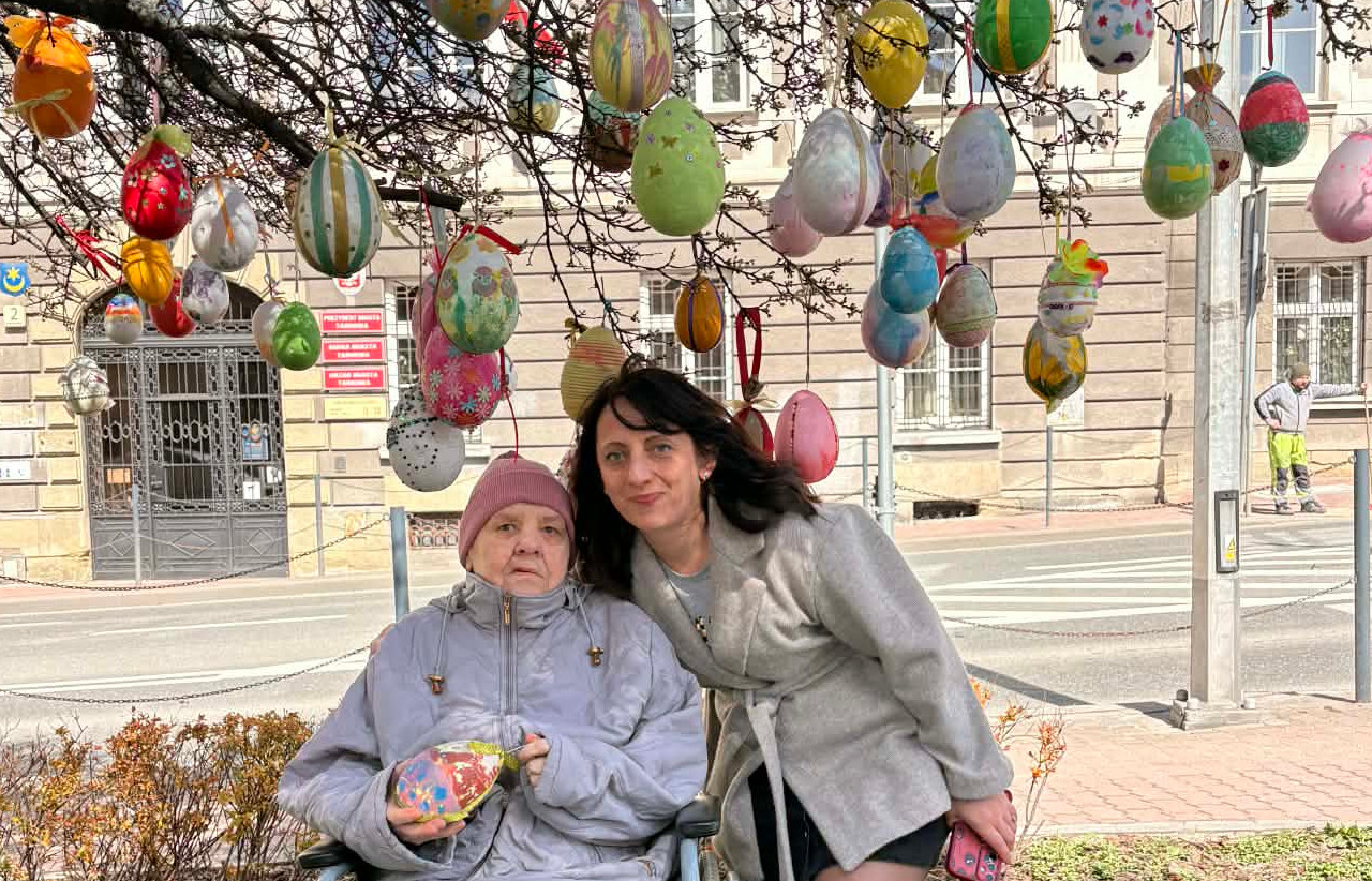 Pracownica Domu oraz Seniorka siedzące na wózku inwalidzkim pozują do zdjęcia pod drzewkiem ozdobionym jajkami wielkanocnymi