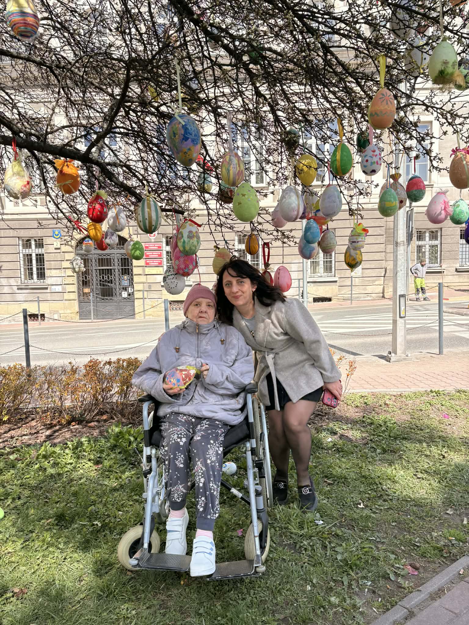 Pracownica Domu oraz Seniorka siedzące na wózku inwalidzkim pozują do zdjęcia pod drzewkiem ozdobionym jajkami wielkanocnymi